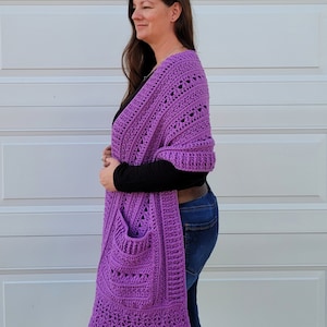 May include: A purple crochet shawl with a large pocket on the front. The shawl is made with a lacy stitch pattern and has a scalloped edge.