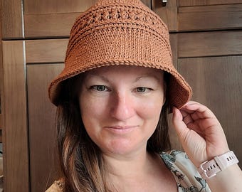 May include: A brown crocheted bucket hat with a textured stitch pattern. The hat is being worn by a person with long brown hair.