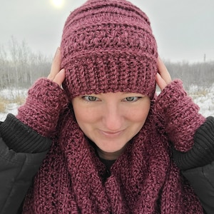May include: A burgundy crocheted hat, scarf, and fingerless gloves set. The hat has a ribbed texture and the scarf and gloves have a textured stitch pattern. The set is photographed against a snowy background.