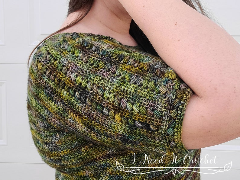 May include: A close-up of a green, brown, and purple crochet top with a short sleeve. The top has a textured stitch pattern and is made with a variegated yarn.