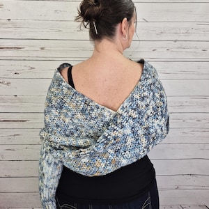 May include: A blue, white, and tan crocheted shawl is draped over the shoulders. The shawl is made of a soft, textured yarn. The person is wearing a black top and blue jeans. The text "I Need It Crochet" is visible on the lower right.