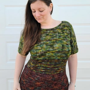 May include: A woman wearing a green, brown, and yellow striped crochet top with short sleeves. The top has a darker brown bottom with a crocheted border. The top is made with a gradient yarn. The woman is wearing blue jeans. The image includes the text "I Need It Crochet".