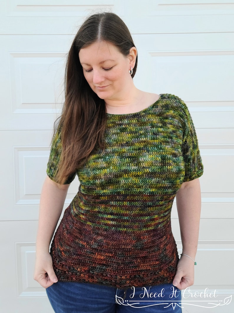 May include: A woman wearing a green, brown, and yellow striped crochet top with short sleeves. The top has a darker brown bottom with a crocheted border. The top is made with a gradient yarn. The woman is wearing blue jeans. The image includes the text "I Need It Crochet".