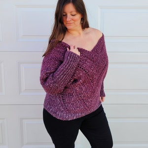May include: A woman wearing a loose-fitting, purple, crocheted sweater with a V-neck and a lacy pattern. She is wearing black pants and has long brown hair.