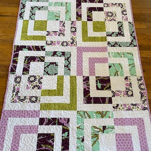 May include: A quilt with a patchwork design featuring squares of purple, green, and white fabric. The quilt is made up of different patterns, including floral, geometric, and abstract designs.