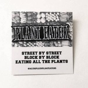 May include: Black and white enamel pin with the text "PLANT EATER" in bold letters. The pin is on a white background with a printed image of a grocery store produce section. The text below the pin reads "STREET BY STREET BLOCK BY BLOCK EATING ALL THE PLANTS" and "@ACOUPLEOFPLANTEATERS".