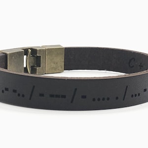 以下が含まれることがあります： 真鍮製の留め具が付いた茶色の革製のブレスレット。ブレスレットには「C+1」という文字が刻印されており、点字でメッセージが書かれています。