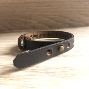 May include: A brown leather bracelet with a brass rivet and the text "king got this! -Code" engraved on the band.