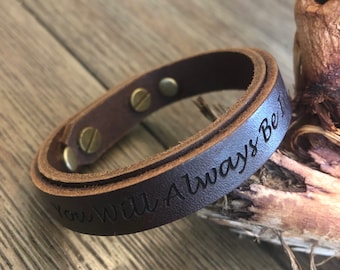 May include: A brown leather bracelet with a silver clasp and the inscription "You Will Always Be" engraved on the band.