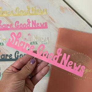 Può includere: Adesivo in vinile rosa glitterato con il testo "Share Good News" in un carattere corsivo. L'adesivo è su uno sfondo trasparente ed è tenuto da una mano.