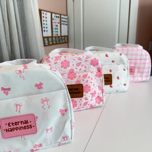 Könnte beinhalten: Vier Lunchpakete mit verschiedenen Mustern: hellblau mit rosa Schleifen, rosa Blumen, weiß mit Erdbeeren und rosa Vichy-Karo. Jede Tasche hat einen weißen Griff und eine Vordertasche. Die erste Tasche hat ein rosa Etikett mit der Aufschrift "Eternal Happiness".