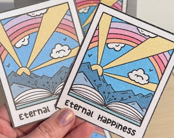 Puede incluir: Tarjetas con una colorida ilustración de un arcoíris, sol, montañas y un libro abierto. El texto "Eternal Happiness" está impreso en las tarjetas. Las tarjetas tienen un borde brillante y se sostienen en una mano.
