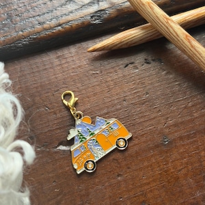 May include: An orange and white enamel charm in the shape of a camper van, with a mountain and tree design on the roof. The charm has a gold-colored clasp and is set against a wooden surface. Knitting needles are in the background.