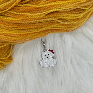 Puede incluir: Un abalorio de metal en forma de oso polar blanco con un gorro de Papá Noel rojo, para un proyecto de hilo o tejido.