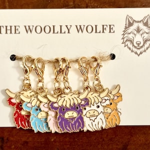 May include: A set of five gold-tone metal lobster clasp charms shaped like Highland cows in red, blue, purple, white, and tan enamel. The charms are attached to a white display card labeled THE WOOLLY WOLFE with a wolf head logo graphic.