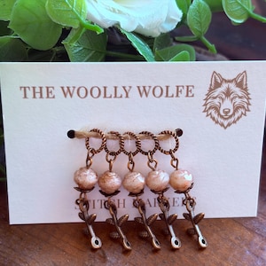 Puede incluir: Cinco marcadores de puntos de color cobre sujetos a una tarjeta blanca con el texto THE WOOLLY WOLFE y un logotipo de lobo. Cada marcador presenta un anillo trenzado, una cuenta redonda de color beige jaspeado y un dije metálico en forma de hoja.