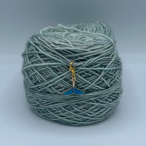 Op de afbeelding: Een blauwe en gouden garenmarker in de vorm van een walvisstaart op een bol lichtblauw garen.
