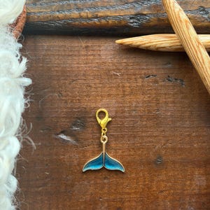 Pode incluir: Um berloque dourado com um design de cauda de baleia azul. O berloque tem um fecho lagosta para fácil fixação. O fundo é uma superfície de madeira com agulhas de tricô e lã branca.
