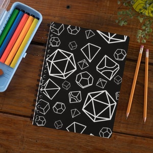 Puede incluir: Una libreta negra de espiral con un patrón geométrico blanco de varias formas, incluyendo cubos, pirámides y dados. La libreta está sobre una mesa de madera con un estuche de lápices azul y dos lápices amarillos.