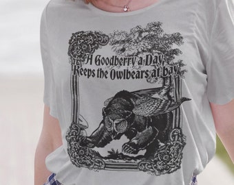 Puede incluir: Una mujer lleva una camiseta gris con un estampado gráfico en blanco y negro de un owlbear. El estampado gráfico incluye el texto "A Goodberry a Day Keeps the Owlbears at Bay". Lleva una camisa de cuadros atada a la cintura y unos vaqueros azules. Lleva una tabla de skate verde.