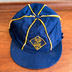 May include: A navy blue baseball cap featuring yellow piping along the seams and a diamond-shaped patch on the front. The patch displays a stylized wolf head graphic above the text CUB SCOUTS in yellow embroidery on a navy blue background.