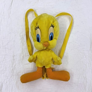 May include: A plush Tweety Bird backpack featuring a bright yellow body, large blue eyes with long black eyelashes, and an orange beak. The character has long orange feet and two yellow fabric straps attached to the back for carrying.