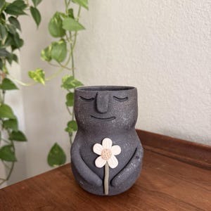 May include: A dark gray, speckled ceramic planter shaped like a stylized figure with closed eyes and a gentle smile. The figure has arms folded across its midsection, holding a single light pink flower with a textured, tan-colored center.