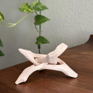 May include: A light-colored wooden display stand composed of two interlocking pieces that form an X-shaped base. The stand features a smooth, natural finish with visible wood grain and carved edges, designed to support spherical objects.
