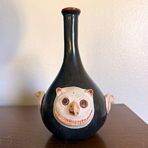 May include: A black ceramic vase with a long, slender neck and a rounded base. A textured, cream-colored face with large eyes, a pointed nose, and a wide, toothy grin is centered on the front. Small, wing-like protrusions extend from the sides.