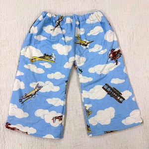 May include: Light blue pants featuring a repeating pattern of various vintage-style airplanes flying among white clouds. The garment has an elasticized waistband and a wide-leg silhouette, showcasing multiple aircraft designs in red, yellow, and brown.