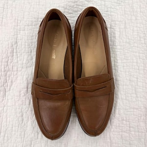 May include: A pair of brown leather loafers featuring a classic penny strap across the vamp, a rounded toe, and a low heel. The interior insoles are tan and clearly display the Clarks Cushion brand logo printed in gold lettering on the heel area.