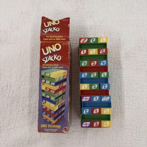 May include: A red cardboard box for the UNO Stacko game featuring the title and a graphic of stacked blocks, positioned next to a vertical tower of colorful plastic blocks in red, blue, green, and yellow, each marked with numbers or symbols.