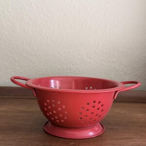 May include: A small, round, red metal colander featuring two side handles and a circular base. The bowl of the colander is perforated with numerous evenly spaced circular drainage holes throughout its body, designed for straining food items.