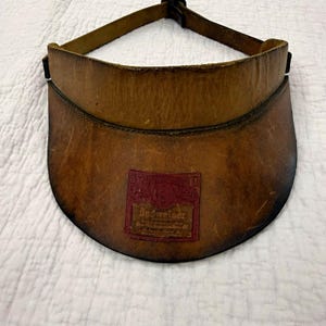 May include: A brown leather sun visor featuring a weathered texture and a rectangular red patch centered on the brim. The patch displays the text Budweiser in embossed lettering. The visor includes an adjustable leather strap at the back.