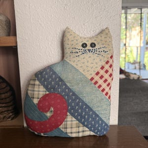 May include: A stuffed cat figure made from various fabric patches in blue, red, cream, and plaid patterns. The cat features two black button eyes and embroidered black whiskers. The fabric includes small floral prints and a red spiral tail design.