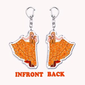 May include: Two clear acrylic keychains with a cartoon illustration of a person in an orange dress holding a microphone. The dress has white floral accents. The keychains have silver-coloured metal hardware. The text "INFRONT BACK" is in orange.