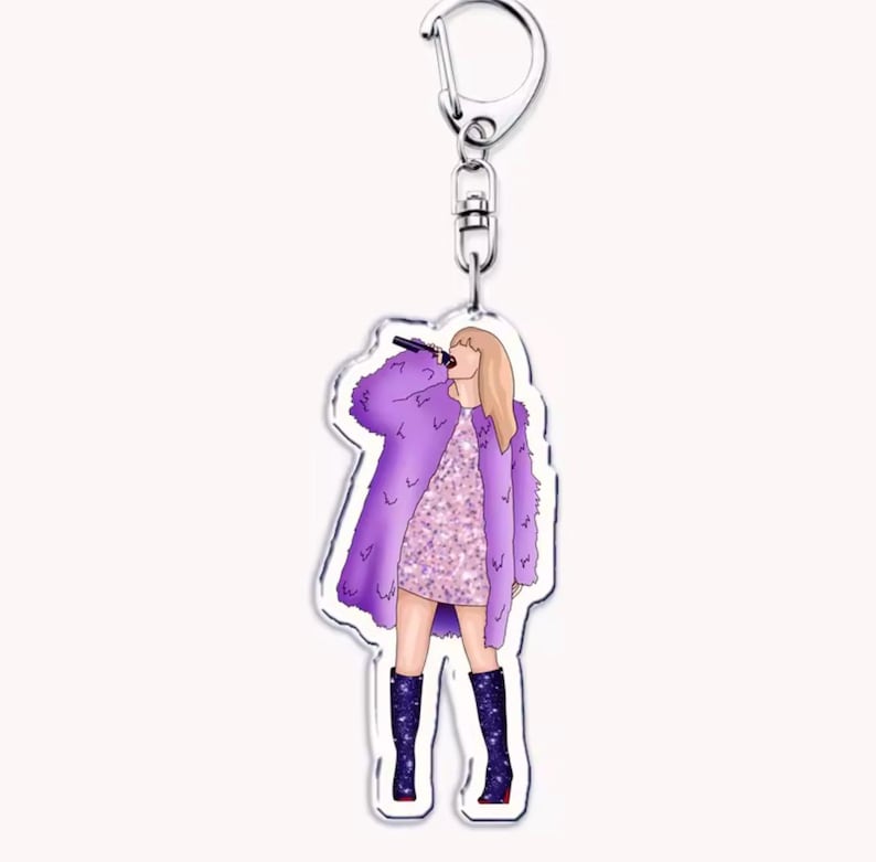 May include: A keychain with a cartoon illustration of a person singing into a microphone. The person is wearing a purple faux fur coat, a sparkly pink dress, and dark blue boots with star accents. The keychain has a silver clasp.