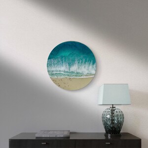 May include: A round wall art piece depicting a beach scene. The artwork features a gradient of colors, from deep teal to white, representing the ocean waves crashing onto a sandy shore. The piece is mounted on a white wall.