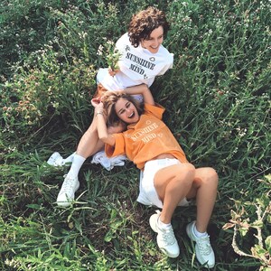 Puede incluir: Dos personas con camisetas a juego con el texto "Sunshine in my mind" impreso en ellas. La persona con la camiseta naranja está tumbada en la hierba y sonriendo. La persona con la camiseta blanca está sentada y mirando a la cámara.