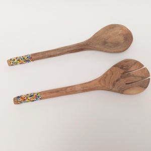 Puede incluir: Dos utensilios de ensalada de madera, una cuchara y un tenedor. Ambos tienen una banda de cuentas de colores en el extremo del mango. La veta de la madera es visible. El fondo es blanco.