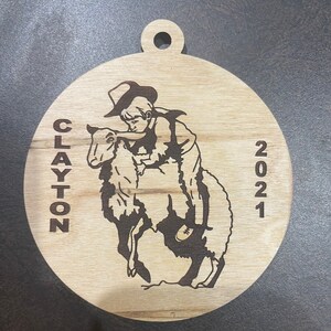 May include: A wooden Christmas ornament featuring a black and white illustration of a boy riding a sheep. The ornament is round and has a hole at the top for hanging. The text "CLAYTON 2021" is engraved on the ornament.
