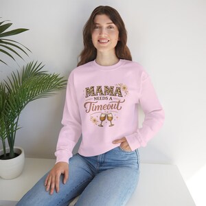 May include: A light pink sweatshirt with the text "MAMA needs a Timeout" in brown and tan lettering. The design includes two wine glasses and floral accents. The long-sleeved top is a casual wear item.