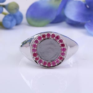 May include: A silver-toned ring with a circular, gray center stone encircled by small, vibrant red gemstones. The ring has a smooth, polished finish and a classic signet style. The background features blue and purple floral accents.