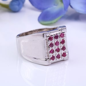 May include: A silver ring with a square face set with a grid of nine small, round, red gemstones. The ring has a polished finish and a wide band. The background includes blue flowers.