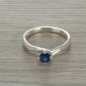 Könnte beinhalten: Ein silberner Ring mit einem einzelnen blauen Saphir-Edelstein, der in einer Krappenfassung sitzt. Die Ringschiene ist gebogen und hat ein schlichtes Design.