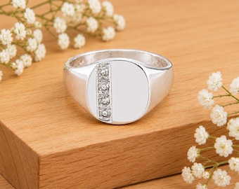 Puede incluir: Un anillo de sello de plata con una cara circular pulida y un canal vertical engastado con cuatro piedras pequeñas, redondas y transparentes. El anillo tiene una banda metálica lisa y reflectante y una sección plana sin adornos.