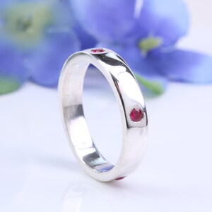 May include: A polished silver ring featuring three small, round, red gemstones. The ring's band is smooth and reflective. A blurred blue flower provides a soft backdrop. The ring's design is simple yet elegant, suitable for everyday wear.
