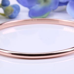 May include: A rose gold bangle bracelet. The smooth, circular bracelet is made of polished metal and reflects light. The background includes blue flowers and a white surface. This jewelry item is a simple, elegant accessory.