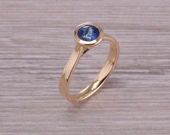 May include: A gold ring with a bezel setting featuring a round blue sapphire gemstone.