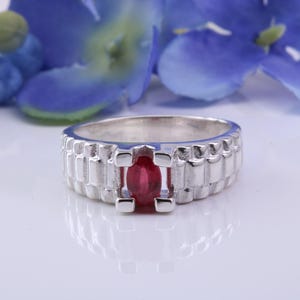 May include: A silver ring with a rectangular design and a central oval-cut ruby gemstone. The ring is set against a white surface with blue flowers in the background, creating a contrast of colors and textures.